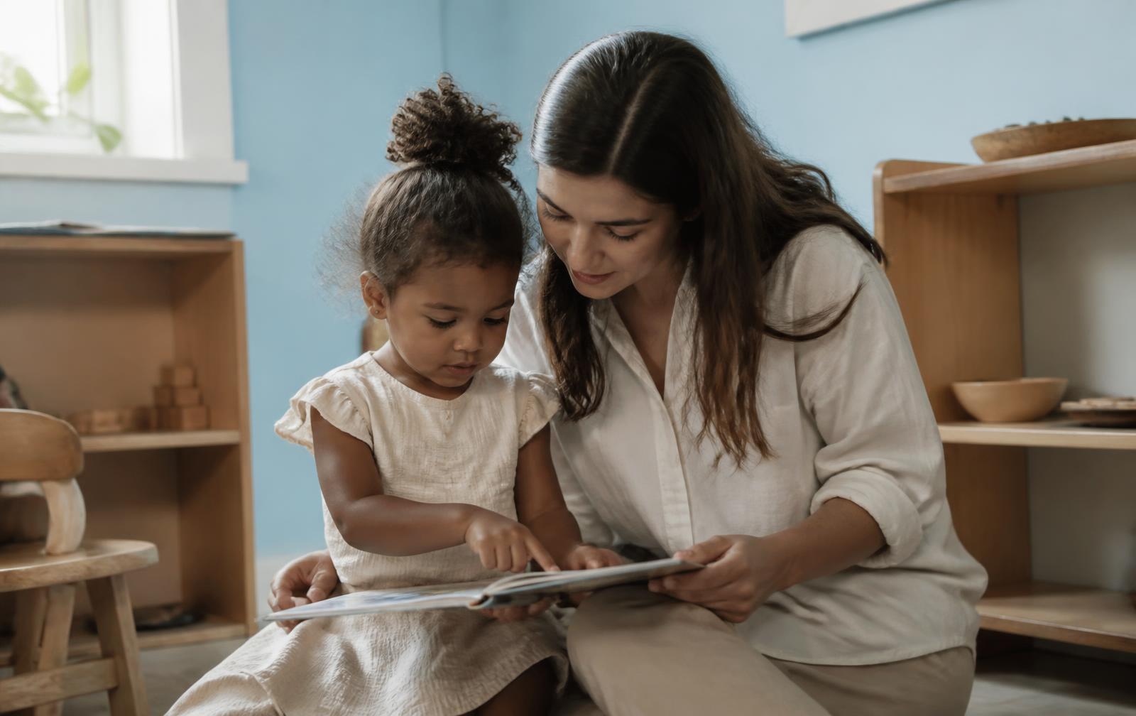 An early childhood educator and a young child sharing a picture book together in a warm, naturally lit playroom