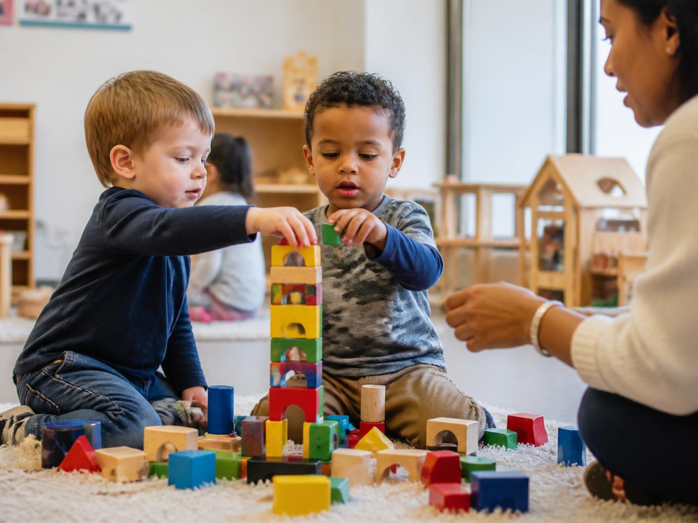 Two young children stacking colorful wooden blocks on a rug with a teacher nearby